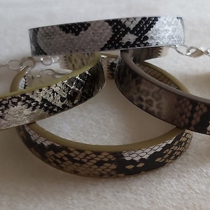 May include: Four snake skin patterned bracelets in shades of brown, grey, and white. The bracelets are stacked on top of each other.