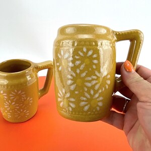 May include: Two mustard-yellow ceramic mugs with white daisy patterns. The mugs have a handle and a cylindrical shape. The larger mug is held by a person. The mugs are on an orange surface with a white background.