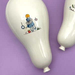 Puede incluir: Dos piezas decorativas de cerámica blanca con forma bulbosa, cada una con una ilustración caprichosa de una persona. Una figura viste un atuendo azul, la otra un atuendo rojo. Las piezas están sobre un fondo morado.