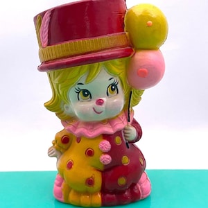 May include: A vintage clown figurine with a red top hat, yellow hair, and a pink ruffled collar. The clown wears a two-toned outfit of yellow and maroon with pink polka dots. It holds two balloons, one yellow and one pink.