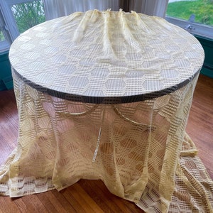 May include: A round table draped with a cream-coloured, patterned lace tablecloth. The tablecloth reaches the floor, and the table has a dark rim and gold-coloured legs. The table is on a hardwood floor.