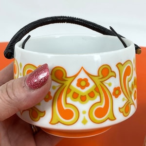 May include: A white ceramic sugar bowl with a black wire handle. The bowl has a geometric pattern in orange and yellow.