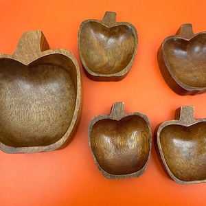 Könnte beinhalten: Ein Set aus fünf holzgeschnitzten Schalen in Apfelform. Die Schalen sind unterschiedlich groß und haben eine natürliche Holzmaserung. Jede Schale hat einen stielartigen Griff. Die Schalen sind auf einer orangefarbenen Oberfläche angeordnet.