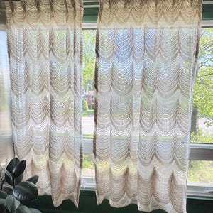 May include: Two sheer, beige curtains with a scalloped design hang in front of a window. The curtains are made of a light, airy fabric, allowing sunlight to filter through. The curtains are hung on a rod and frame a view of the outdoors.