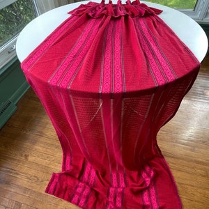 Peut inclure: Un textile rouge drapé sur une table blanche. Le textile a un bord à volants et un motif à rayures avec des accents géométriques. Le tissu est rouge foncé avec des détails roses plus clairs.