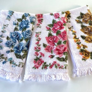 May include: Three white hand towels with fringed edges. Each towel features a different floral pattern: blue roses, pink roses, and yellow roses.