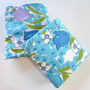 May include: Two folded pieces of fabric featuring a white background with a repeating floral pattern in shades of light blue, medium blue, lavender, and olive green. The design includes stylized tulips and small blossoms with green leaves.