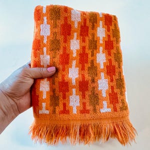 May include: A rectangular orange hand towel featuring a repeating geometric pattern of vertical columns in shades of white, light orange, and dark brown. The towel is finished with a fringed edge along the bottom, showcasing a soft, textured fabric.