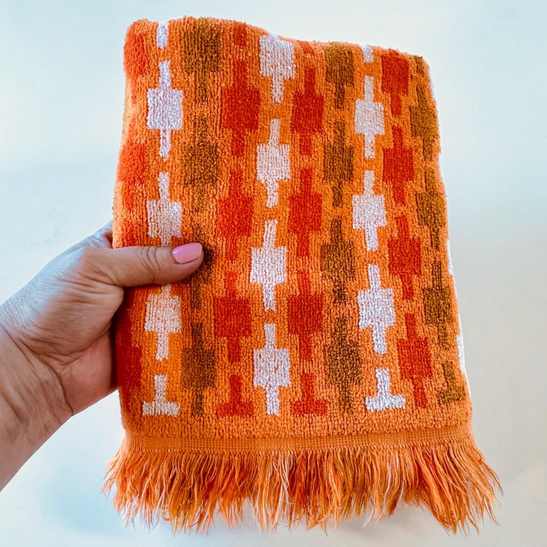 May include: A rectangular orange hand towel featuring a repeating geometric pattern of vertical columns in shades of white, light orange, and dark brown. The towel is finished with a fringed edge along the bottom, showcasing a soft, textured fabric.