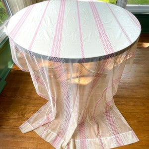 May include: A round table draped with a white tablecloth with pink vertical stripes. A sheer, cream-coloured overlay with pink trim hangs over the table. The table has a metal frame and is set on a wooden floor. The overall effect is layered.