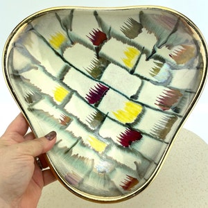 May include: A heart-shaped ceramic serving dish with a gold rim. The dish features a cream-colored background with a woven pattern of yellow, burgundy, and gray brushstrokes. The dish is held by a hand.