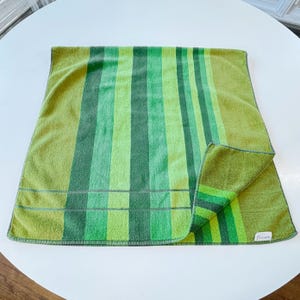 May include: A rectangular terry cloth towel featuring a vertical striped pattern in various shades of lime, olive, and forest green. The towel has a folded corner revealing the underside and a small white rectangular fabric tag on the bottom right edge.