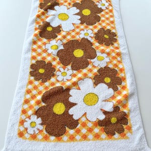 May include: White rectangular towel with a floral pattern. The towel features large brown and white flowers with yellow centers, set against an orange and white checkered background. The edges are white with fringe.
