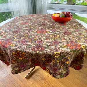 May include: A round tablecloth featuring a repeating pattern of orange, yellow, and green floral motifs enclosed in brown circular outlines. A red bowl containing various fruits sits on the surface of the fabric, which drapes over a table.
