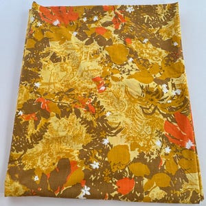 May include: A folded fabric with a floral pattern in shades of yellow, brown, and orange. The design features stylized leaves and flowers, with small white accents. The fabric has a vintage aesthetic.