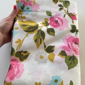 May include: Folded white fabric with a floral pattern featuring pink roses, blue flowers, and green and gold leaves. The fabric is held by a hand with red nail polish.