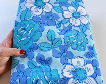 May include: Blue and white floral fabric with a large-scale pattern. The fabric features a variety of flowers, including roses, daisies, and other blooms. The background is a light blue color.
