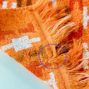 May include: A textured orange towel featuring a geometric pattern in white and dark brown. The fabric has a looped pile construction and a fringed edge. A blue circle highlights a small area of discoloration on the orange fabric near the fringe.