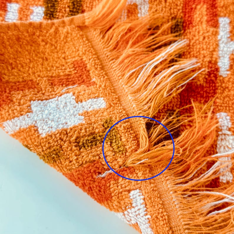May include: A textured orange towel featuring a geometric pattern in white and dark brown. The fabric has a looped pile construction and a fringed edge. A blue circle highlights a small area of discoloration on the orange fabric near the fringe.