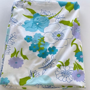 May include: White fabric with a floral pattern featuring large turquoise, light blue, and lavender flowers with green leaves. The flowers are outlined in dark blue, creating a vintage aesthetic. The fabric is folded.