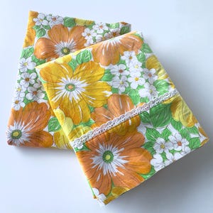 Puede incluir: Dos fundas de almohada plegadas con un vibrante estampado floral de grandes flores naranjas y amarillas, pequeñas flores blancas y hojas verdes. Un delicado borde de encaje blanco está cosido horizontalmente en la funda.