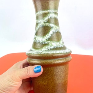 Puede incluir: Un jarrón de cerámica marrón con la parte superior acampanada y una base ancha. El jarrón presenta un patrón de remolino blanco y verde alrededor del centro. El jarrón está sostenido por una persona. El jarrón está sobre una superficie roja.