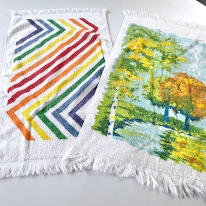Puede incluir: Dos toallas de algodón blanco. Una presenta un diseño de chevron arcoíris. La otra representa un paisaje con árboles y agua. Ambas toallas tienen bordes con flecos.
