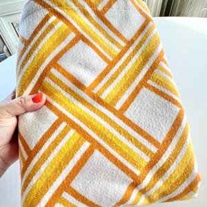 May include: A folded white blanket with a geometric pattern of yellow and orange stripes. The blanket has a soft, textured appearance and is folded neatly. The design features diagonal lines and square shapes.