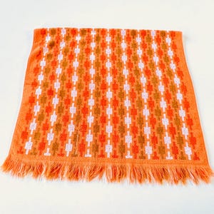 May include: A rectangular orange hand towel featuring a repeating geometric pattern of vertical columns in white and darker orange. The towel has a textured terry cloth surface and is finished with a fringe along the bottom edge.