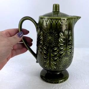 May include: A green ceramic jug with a lid, handle, and pedestal base. The jug is decorated with a floral pattern. The jug is held by a person.