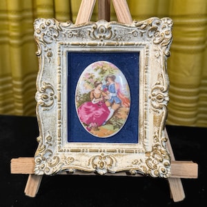 Puede incluir: Un marco ovalado de porcelana que representa a una pareja en un jardín. El marco es ornamentado, con un acabado blanco y dorado envejecido. La placa está colocada sobre un fondo azul y se exhibe en un pequeño caballete de madera.