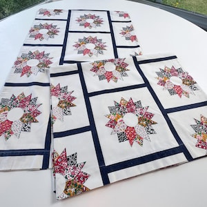 May include: White fabric table runner and placemats with a floral starburst design. The starbursts are made of colorful fabric patches, and the pieces are outlined with navy blue borders. The placemats and runner are on a white surface.