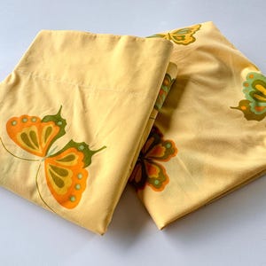 Könnte beinhalten: Zwei gelbe Kissenbezüge mit einem Schmetterlingsdesign. Die Schmetterlinge haben orange, grüne und gelbe Flügel. Die Kissenbezüge sind gefaltet und aufeinander gestapelt.