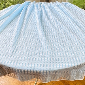 May include: A round table draped with a pale blue tablecloth. The cloth features a textured pattern of white and blue diamond shapes. The table is positioned in front of a window, with a view of greenery.