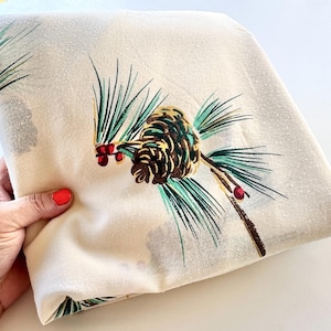 May include: A folded off-white fabric featuring a repeating print of brown pinecones, green pine needles, and small red berries. The textile has a soft, matte texture and displays a botanical pattern across the visible surface of the material.