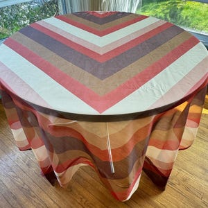 May include: A round table covered with a retro-style tablecloth. The tablecloth features a chevron pattern in shades of red, brown, beige, and white. The table is set in front of a window, and the tablecloth hangs down to the floor.