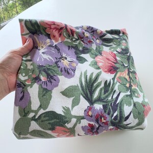 May include: A folded piece of fabric featuring a floral pattern with purple, pink, and green flowers and leaves on a white background. The textile displays a woven texture with a repeating botanical design of various blossoms and foliage.
