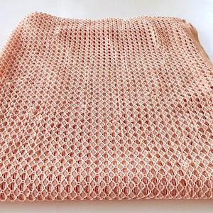 May include: A peach-colored, open-weave fabric, possibly a scarf or shawl, is displayed on a white surface. The fabric has a repeating diamond pattern with small holes throughout, creating a delicate, airy texture. The edges are neatly finished.