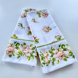 May include: Two white floral print towels. The towels feature a repeating pattern of peach-colored roses and green leaves. The edges are trimmed with a green scalloped border. The towels are folded and displayed on a white surface.