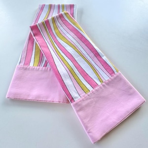 May include: Two pillowcases with a pink and white striped pattern on the top portion and solid pink on the bottom. The stripes include pink, white, and yellow. The pillowcases are made of a soft fabric.