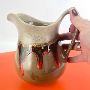 Puede incluir: Una jarra de cerámica con la parte superior beige y la base marrón. La jarra tiene un diseño único con gotas marrones y rojas. El asa es curva y la jarra está sostenida por una mano.