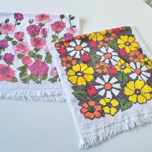 May include: Two rectangular terry cloth towels with fringed edges. One towel features a white background with pink and purple rose patterns. The second towel has a white background with a dense pattern of yellow, orange, and white daisy flowers.