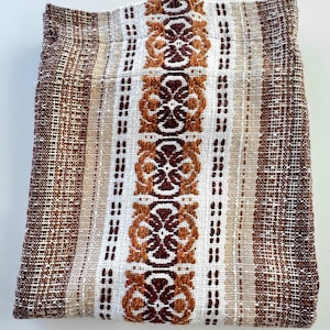 Peut inclure: Un textile tissé avec un motif floral répété dans des tons de marron, beige et blanc. Le tissu a un aspect texturé avec des rayures verticales et un motif central détaillé. Le textile est plié.