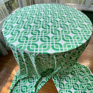 May include: A round tablecloth with a repeating geometric pattern in shades of green and white. The tablecloth is draped over a round table, with the fabric cascading to the floor. The design features interlocking shapes.