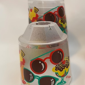 May include: Two clear plastic containers with lids. The containers are decorated with colourful images of sunglasses and ice cream sundaes. The design includes red, green, yellow, and brown colours. The containers are cylindrical.