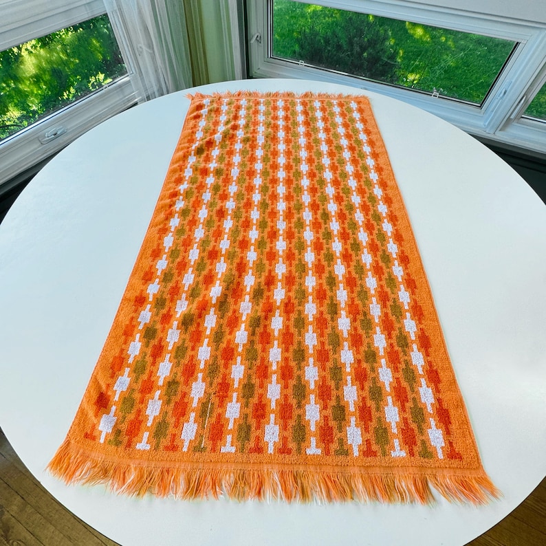 May include: A rectangular orange hand towel featuring a repeating geometric pattern in white and olive green. The towel is constructed from a textured terry cloth fabric and is finished with long, matching orange fringe along both short ends.