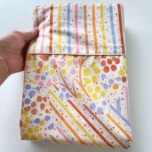 May include: A folded textile piece featuring a cream-colored background with a top section of vertical stripes in pink, orange, yellow, and blue. The lower section displays an abstract pattern of orange, yellow, and blue dots and diagonal lines.