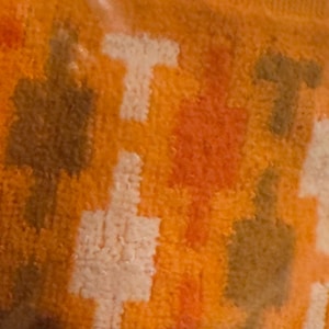 May include: A textured fabric featuring a repeating geometric pattern of T-shaped motifs in shades of white, dark brown, and orange. The surface has a soft, looped pile construction with a consistent density across the entire visible area of the textile.
