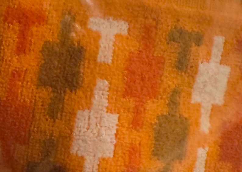 May include: A textured fabric featuring a repeating geometric pattern of T-shaped motifs in shades of white, dark brown, and orange. The surface has a soft, looped pile construction with a consistent density across the entire visible area of the textile.
