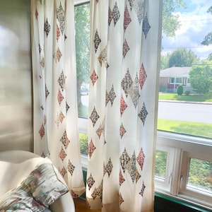 May include: Off-white curtains featuring a diamond design in brown, red, and grey. The curtains are hanging in front of a window, with a view of the outside visible. A white chair and a patterned cushion are in the foreground.